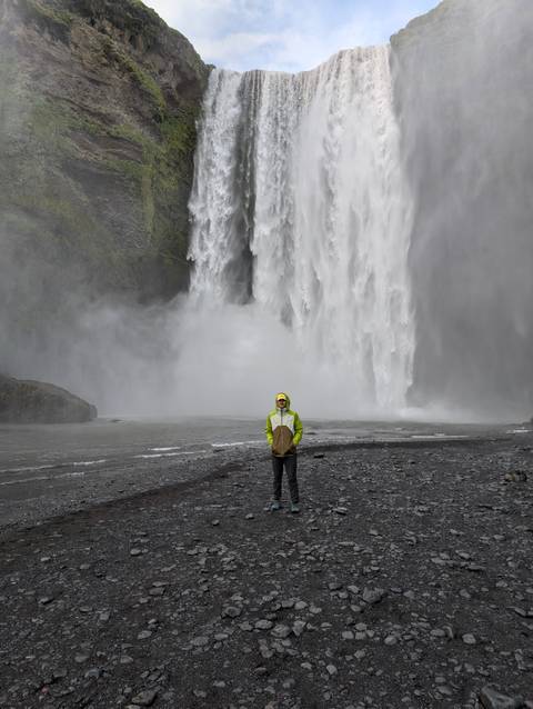 Person standing in front of a large waterfall in a rugged landscape.