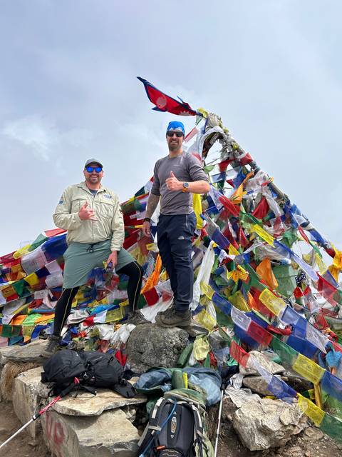 Two men posing with prayer flags at a high altitude.