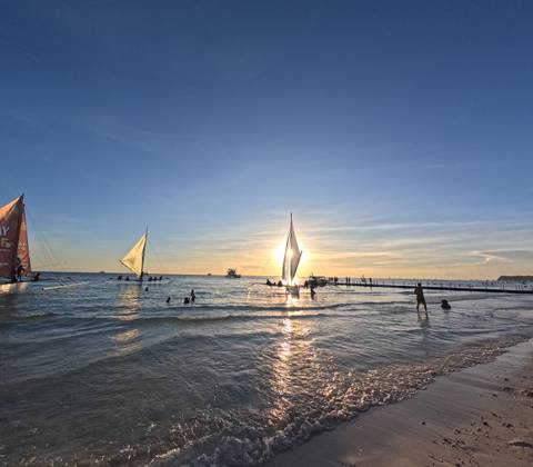       Sailboats on the ocean during sunset with people swimming.
  