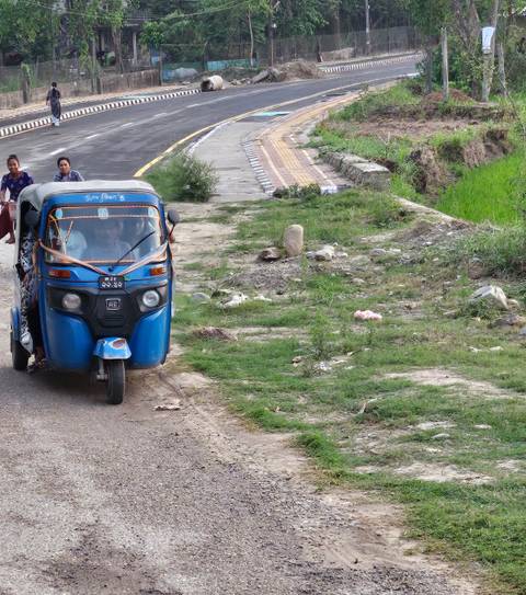       Three-wheeled auto rickshaw driving on a dirt path with people inside.
  
