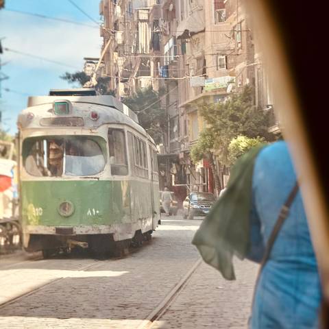       A tram on a street with people walking in the background and a person partially visible in a vehicle.
  
