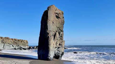 Tall rock formation on a coastal beach with waves.