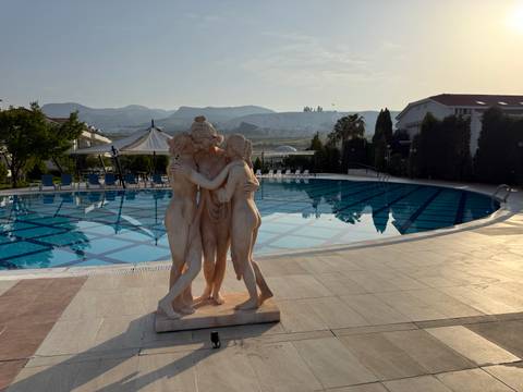       Statue of three women in front of a swimming pool with mountainous background.
  