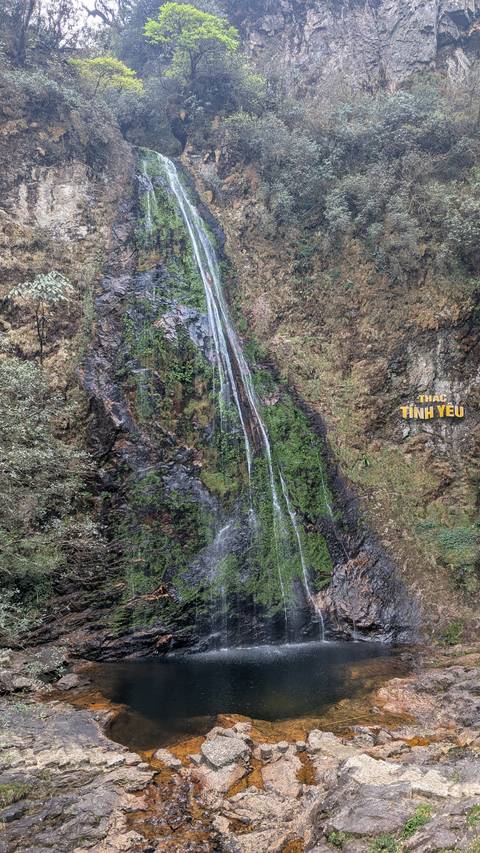       A waterfall flowing down a rocky cliff, surrounded by greenery.
  