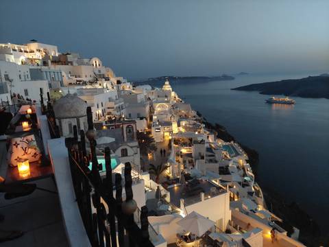       A picturesque view of a coastal village with white buildings lit up at night, overlooking a body of water and a distant cruise ship.
  
