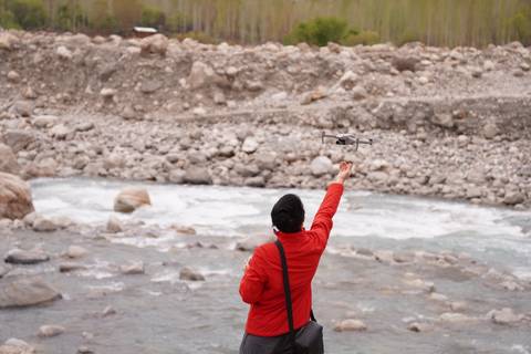       Person standing by a river with a drone, rocky terrain visible.
  