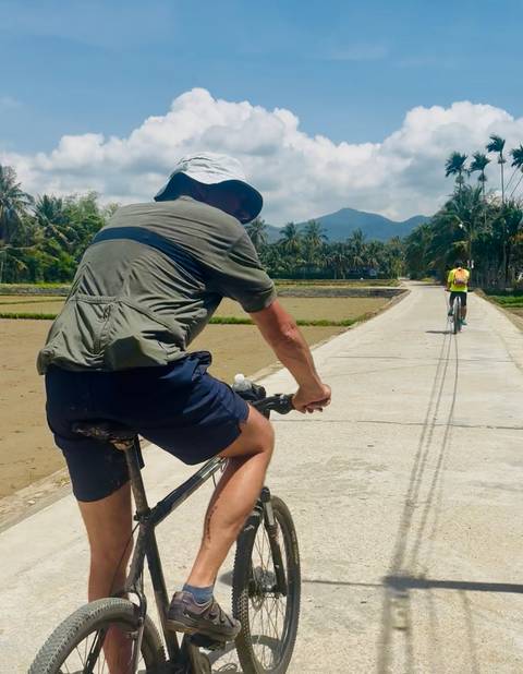       Two cyclists riding along a scenic road surrounded by palm trees and mountains.
  