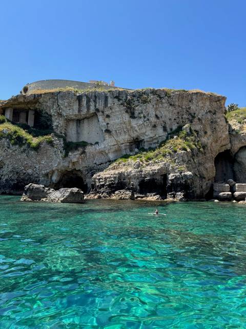 Person swimming near rocky cliffs in clear, turquoise water.