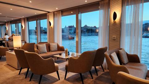 Elegant interior of a cruise or hotel lounge with a view of the river and historic architecture in the background.