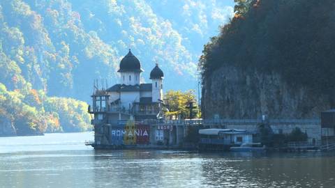 Scenic view of a church by a river with lush hills in the background, and a sign reading 'ROMANIA'.