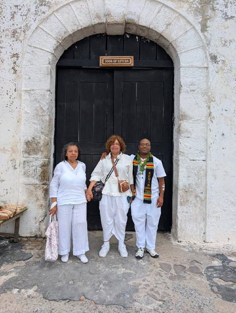 Three people in white clothing posing in front of a large, black wooden door.