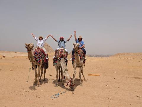 Three people riding camels in the desert with pyramids in the background.