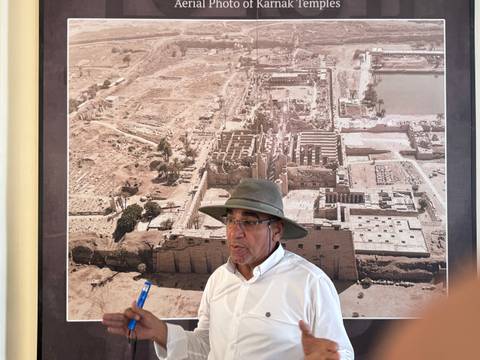       A person in a hat is standing in front of an aerial photograph of an ancient site.
  