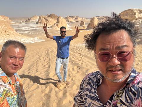       Three people are posing in a sandy desert landscape with rock formations.
  