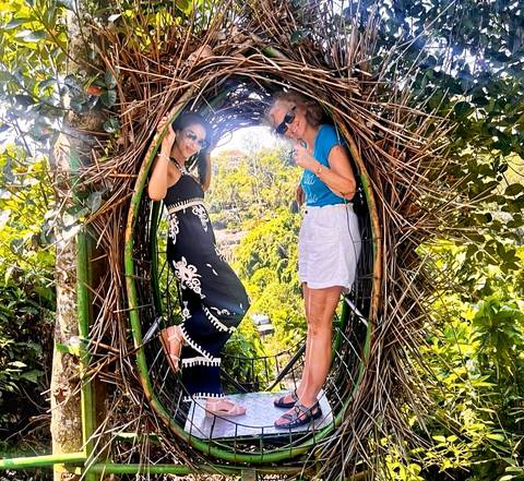       Two women posing inside a decorative nest structure in a lush area.
  