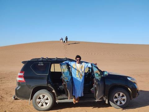       Man standing near a 4x4 in desert dunes with others in the background.
  