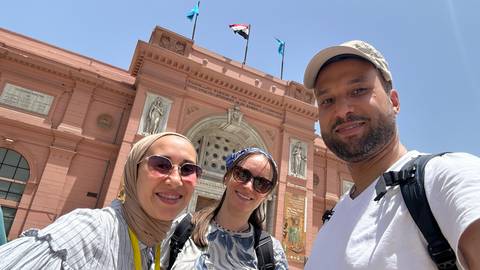 A group of people posing in front of a red historic building with flags.