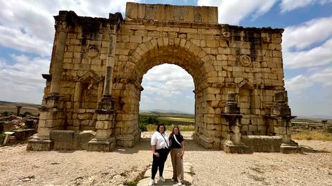       Two people standing under an ancient stone archway.
  