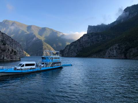Blue ferry carrying a van on a scenic lake surrounded by mountains.