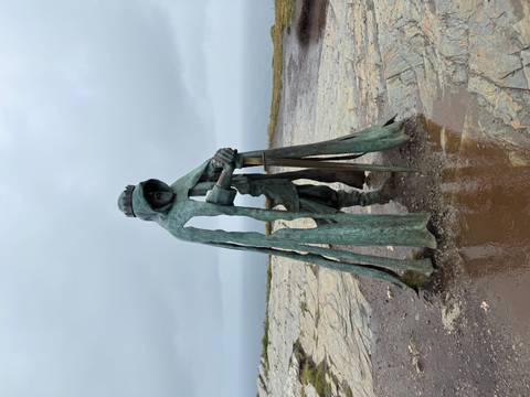 Statue of a robed figure holding a sword with a coastal backdrop.