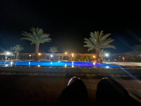 Night view of a lit swimming pool surrounded by palm trees and a building.