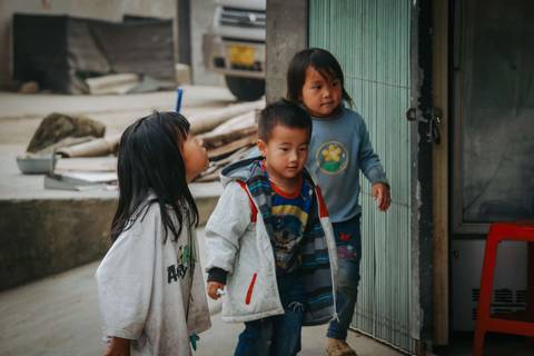       Three children in casual clothing, standing near a building.
  