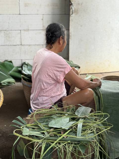       A person sitting cross-legged working with banana leaves indoors.
  