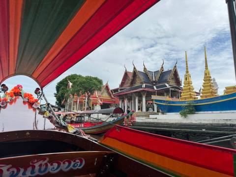       Colorful longtail boat passing by ornate structures by the river.
  