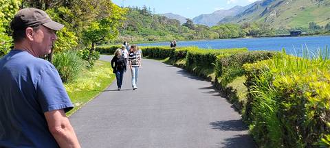       People walking along a lakeside path with mountains in the background.
  