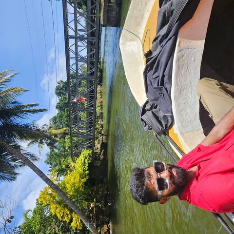 Person in a boat on a river with a metal bridge and palm trees.