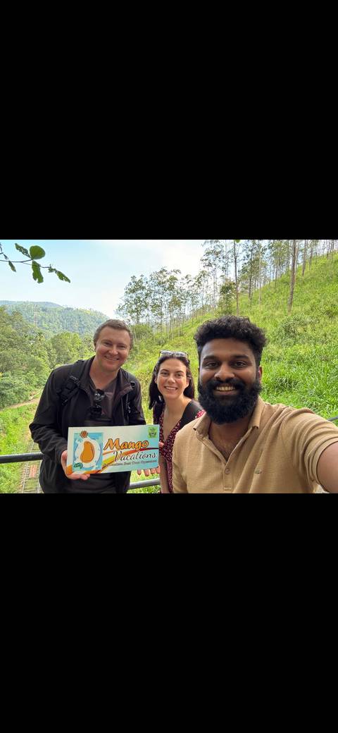 Selfie with three people holding a sign, lush greenery in the background.