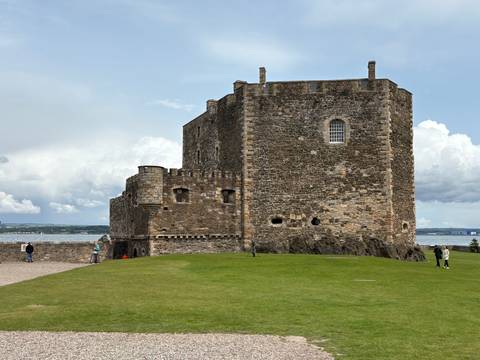 A historic stone castle overlooking a body of water, with grassy grounds and people walking nearby.