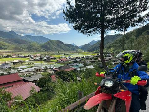 A person on a motorcycle overlooking a valley filled with houses and fields.