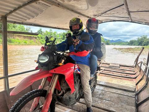       Motorcyclists on a boat crossing a river, open landscape in the background.
  