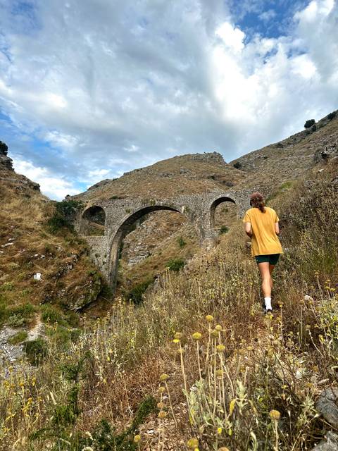       Person walking towards a stone arched bridge in a hilly landscape.
  