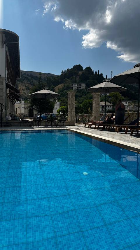       People relaxing by a swimming pool with a view of buildings and trees.
  
