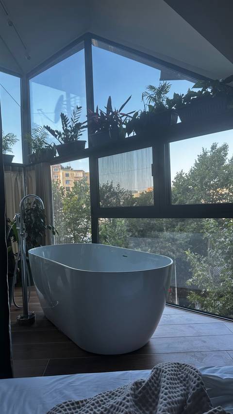 Bathtub by a large window with a view of trees and buildings.