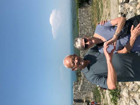 An elderly couple making a heart shape with their hands, with a scenic view in the background.