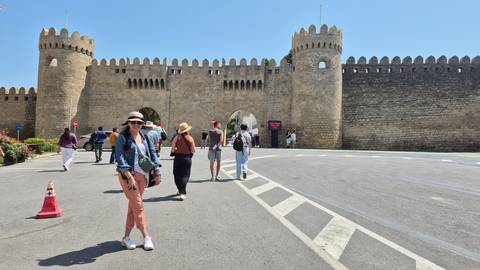       People walking towards a historic stone wall with towers.
  