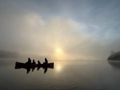       Canoe with people at sunrise on a foggy lake.
  