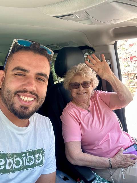       Two people smiling inside a vehicle with greenery visible through the window.
  