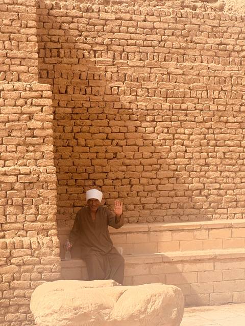       Man in traditional attire waving in front of a brick wall.
  