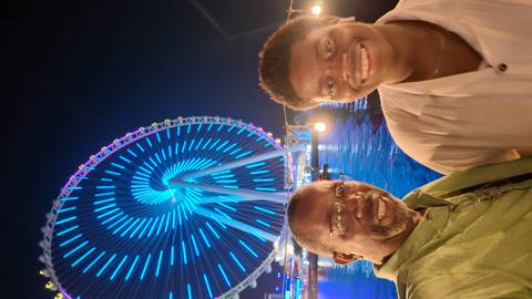 Two people posing in front of a lit Ferris wheel at night.