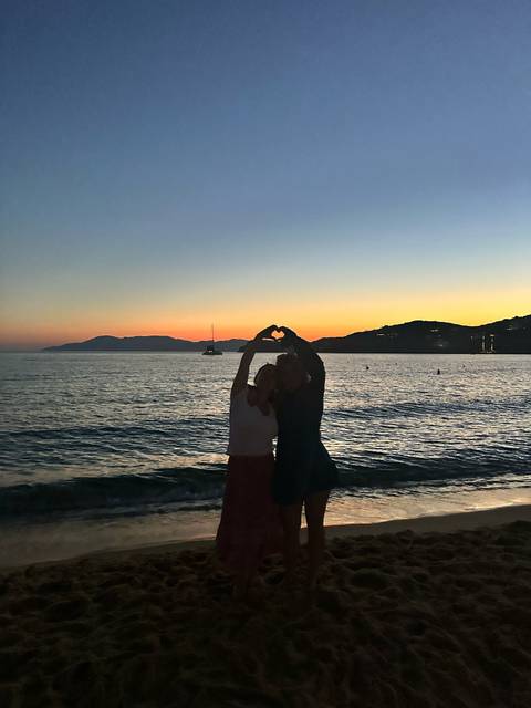 Two people on a beach at sunset forming a heart shape with their hands.