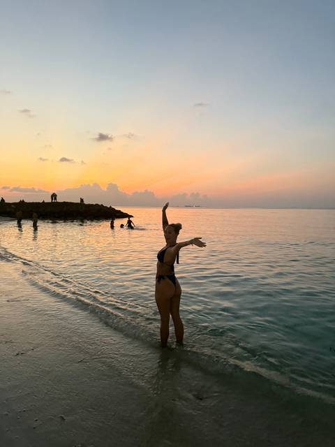       Person standing in the ocean at sunset, posing in swimwear.
  