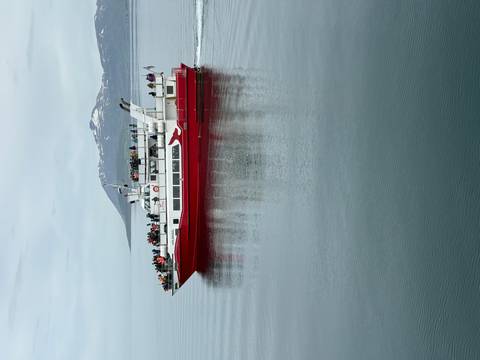 Red boat with people sailing on a calm sea with snowy mountains.