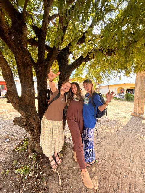       Three women posing joyfully under a tree in a rustic setting.
  