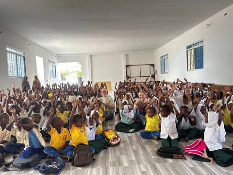Large group of children and a few adults inside a hall, cheering joyfully.