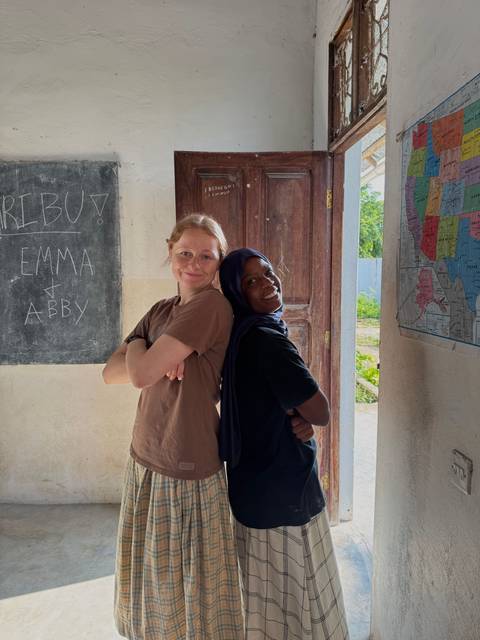       Two women leaning back-to-back inside a building with a map and chalkboard.
  