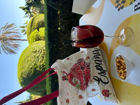       A glass of sangria with a decorative bag and a bowl of snacks on a table, with greenery in the background.
  